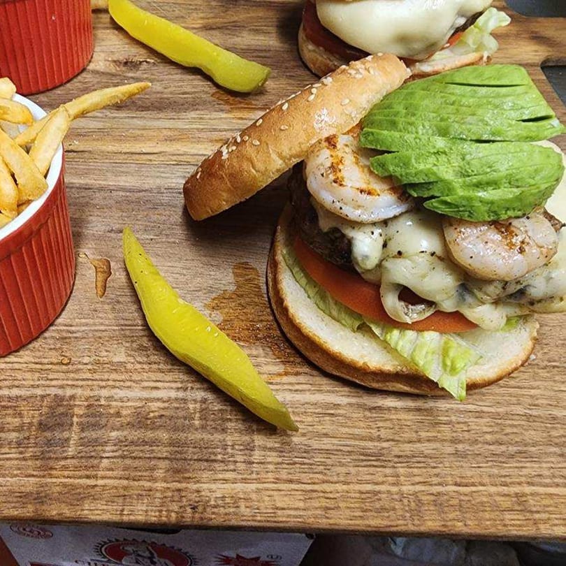 A shrimp and avocado burger with fries and pickled peppers on a wooden board.